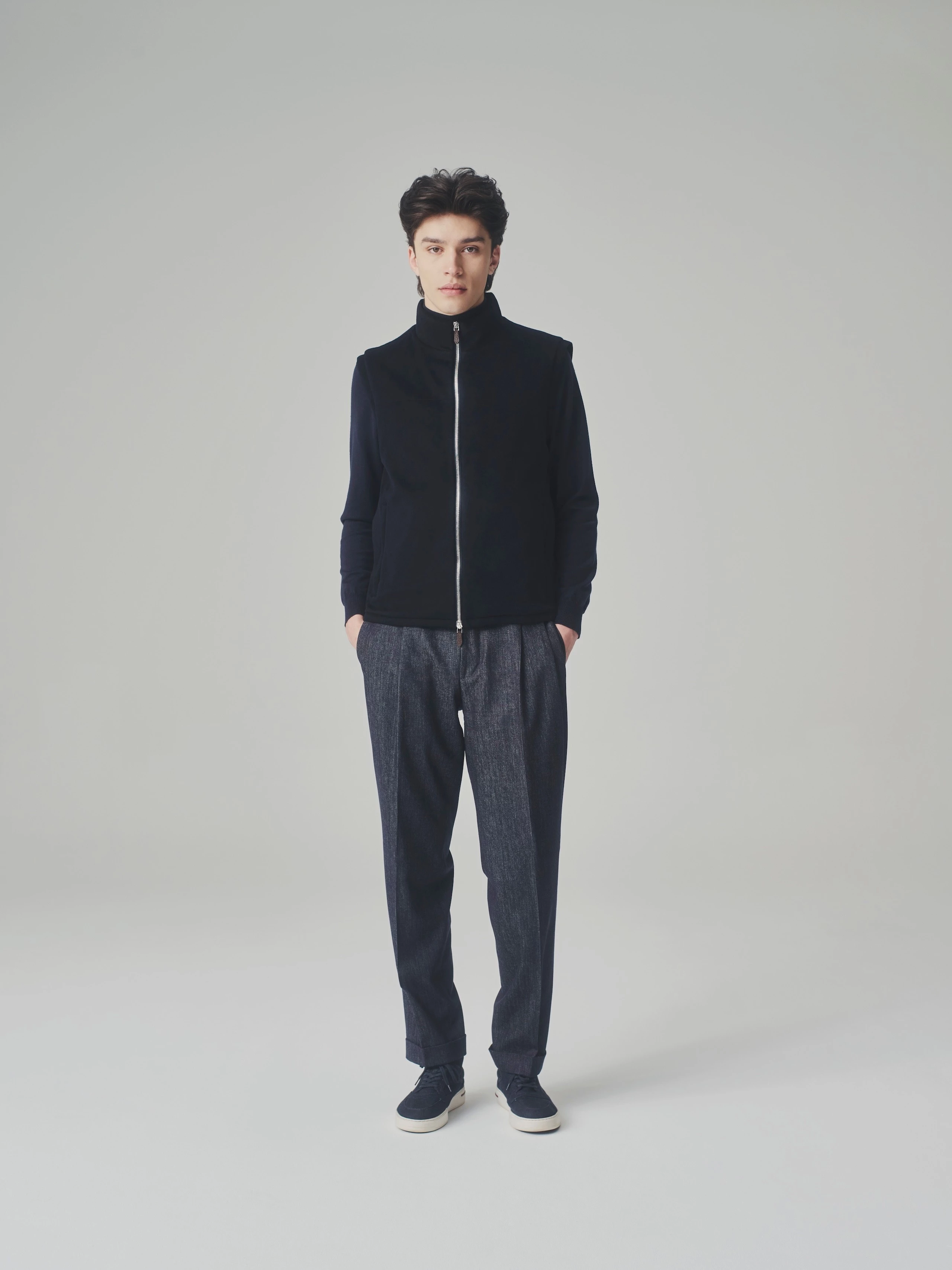 Man in a studio wearing a closed dark blue cashmere bulletproof vest over a cashmere sweater and pleated trousers.