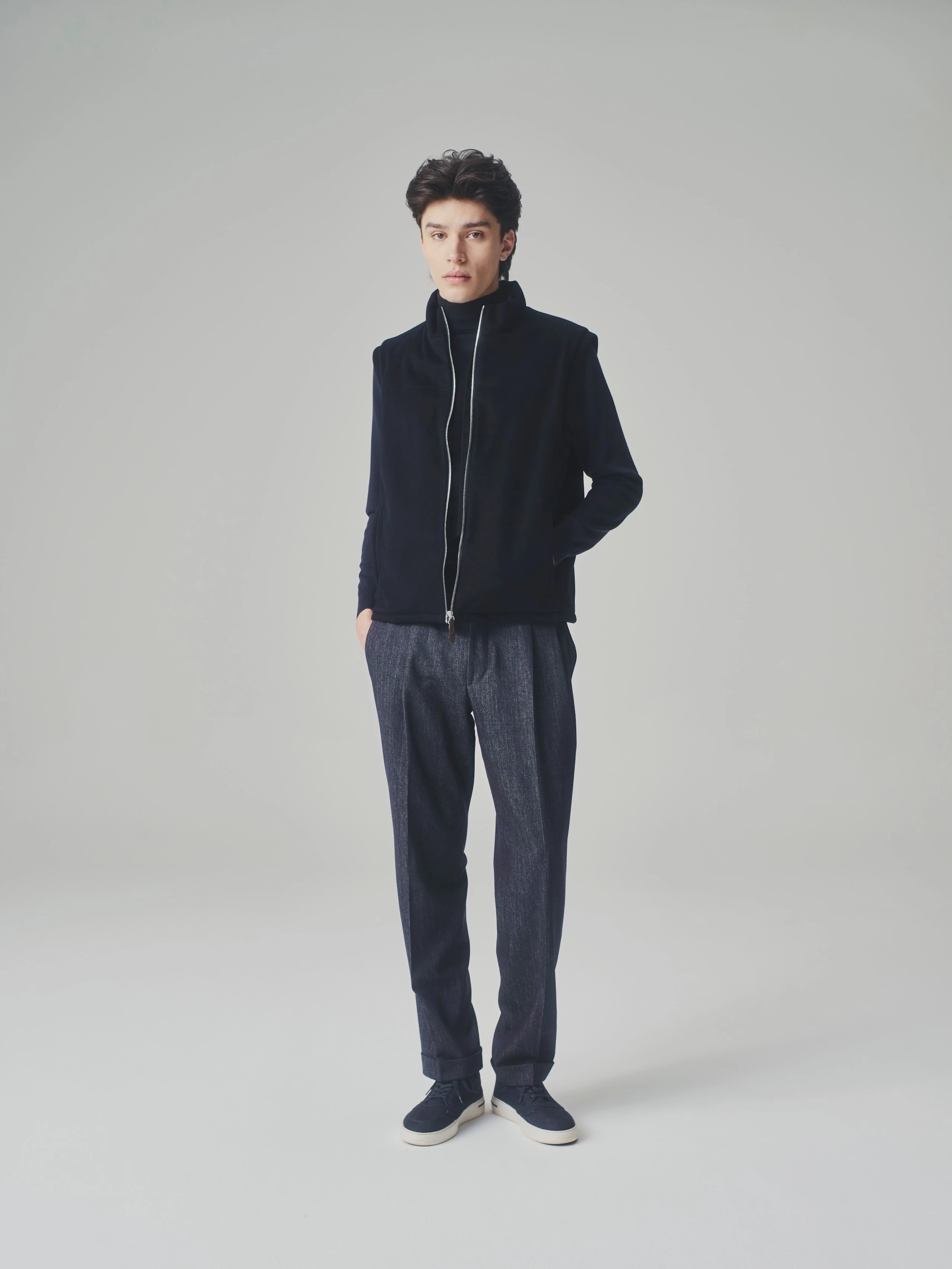 Man in a studio wearing an open dark blue cashmere bulletproof vest, cashmere sweater and pleated trousers.