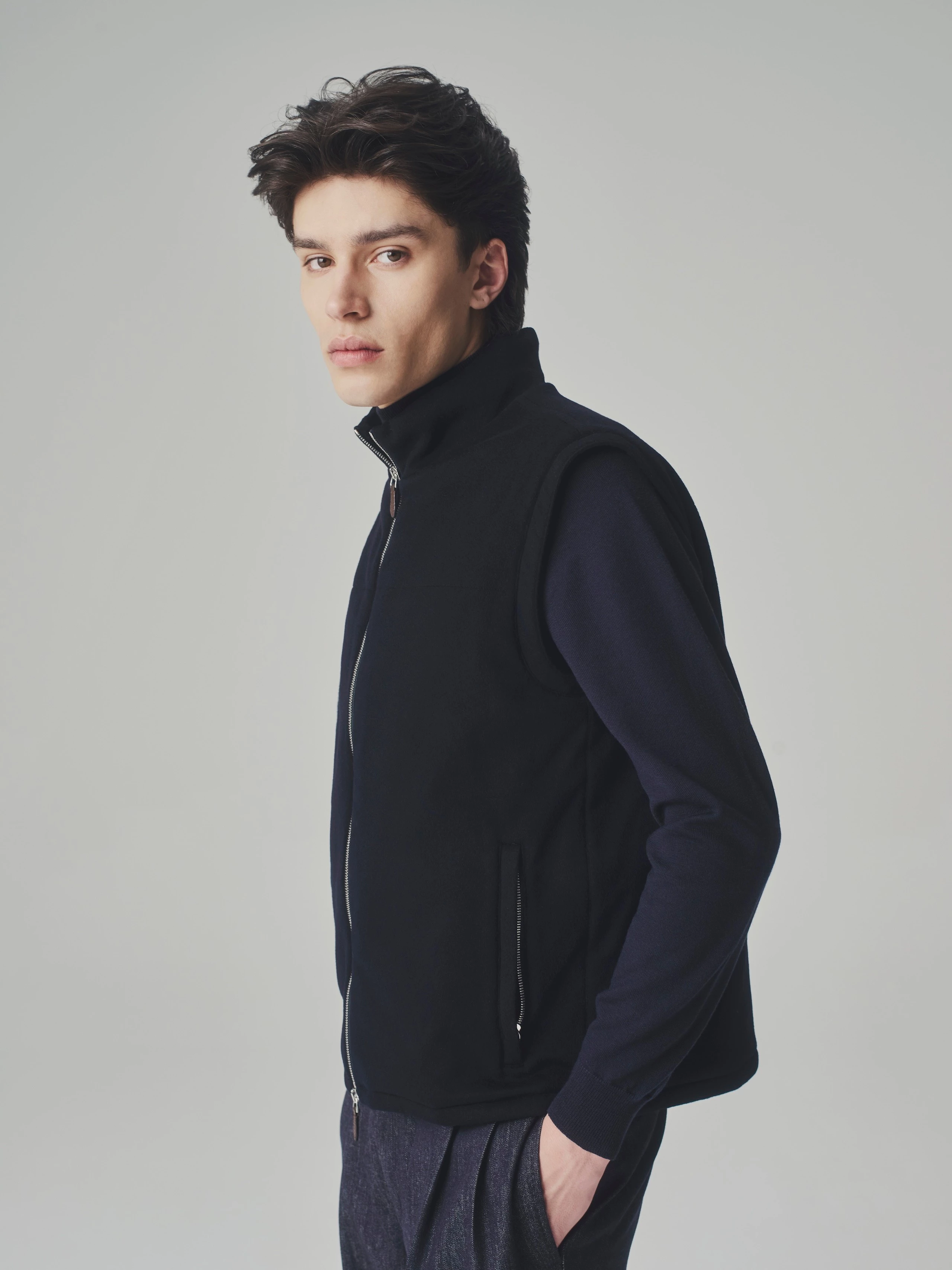 Portrait of a man in a studio wearing a dark blue cashmere bulletproof vest over a cashmere sweater and pleated trousers.