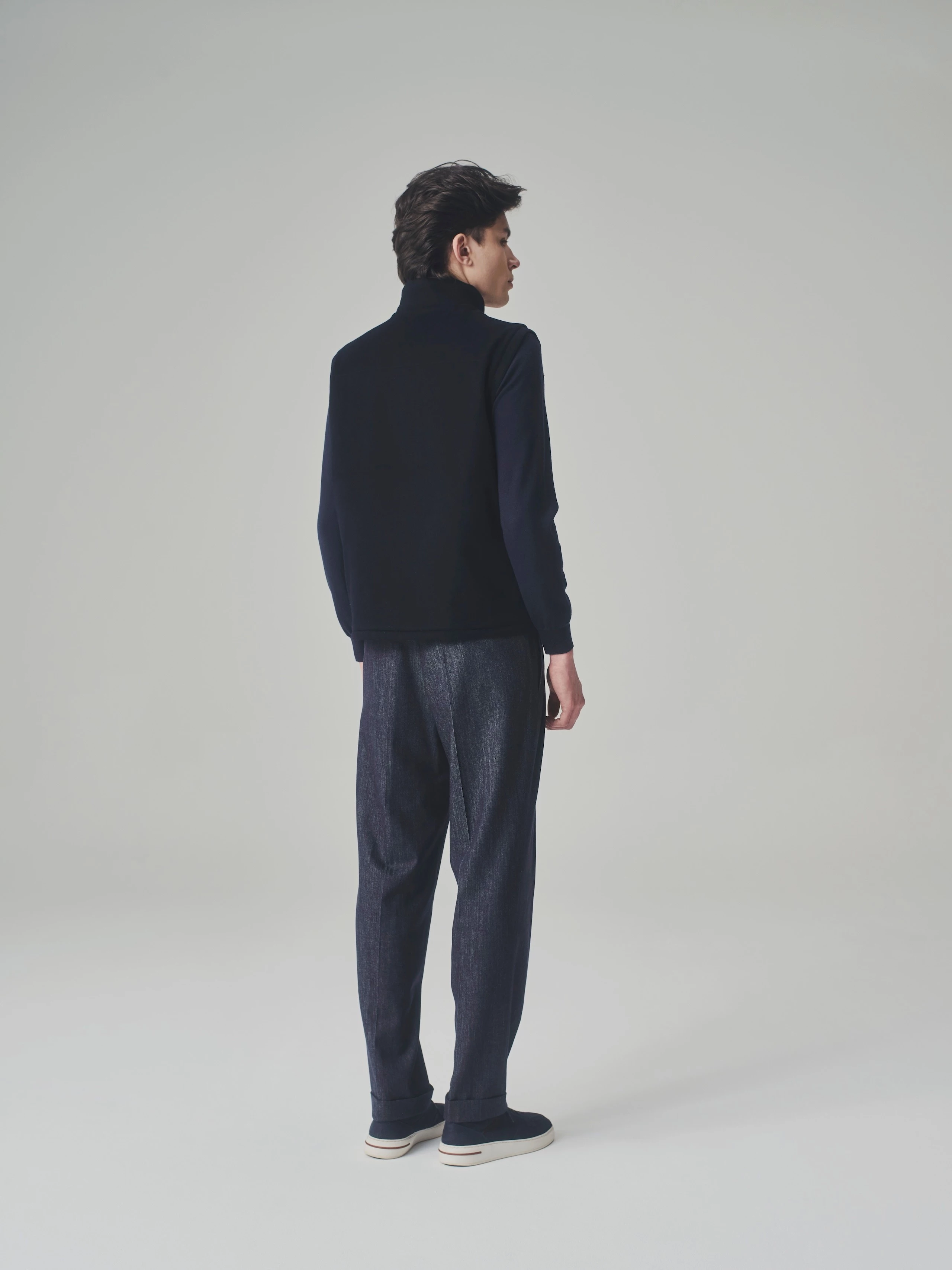 Back view of a man in a studio wearing a dark blue cashmere bulletproof vest, cashmere sweater and pleated trousers.