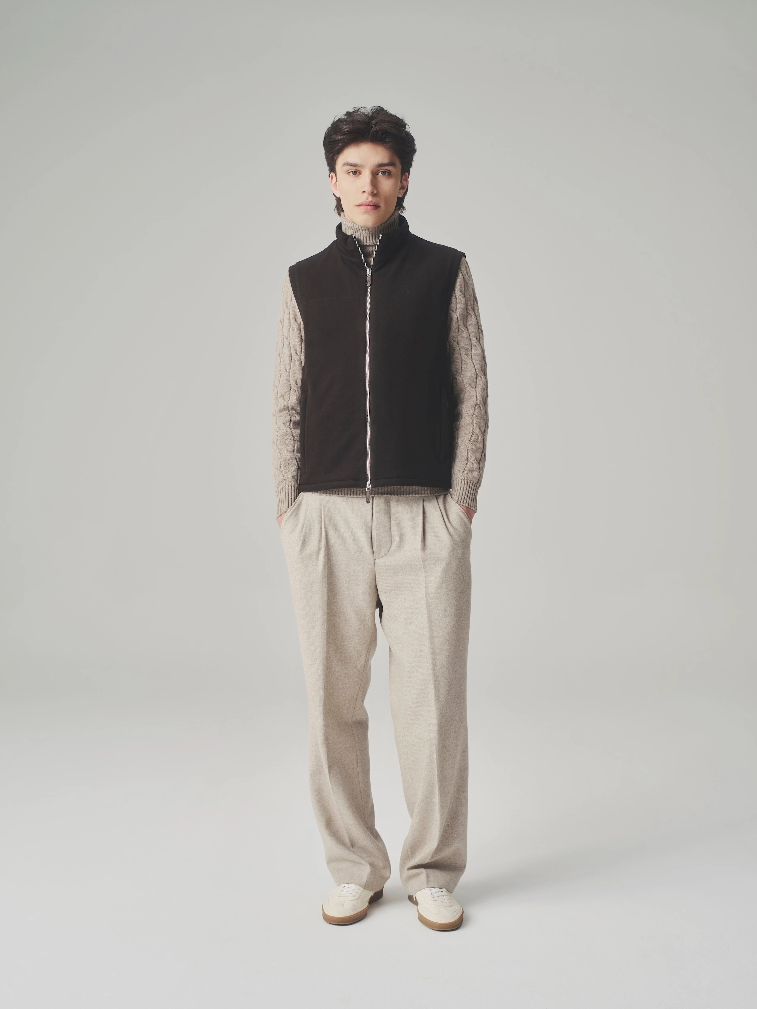 Man standing in a studio wearing a closed brown bulletproof vest over a beige cable knit sweater and wool trousers.
