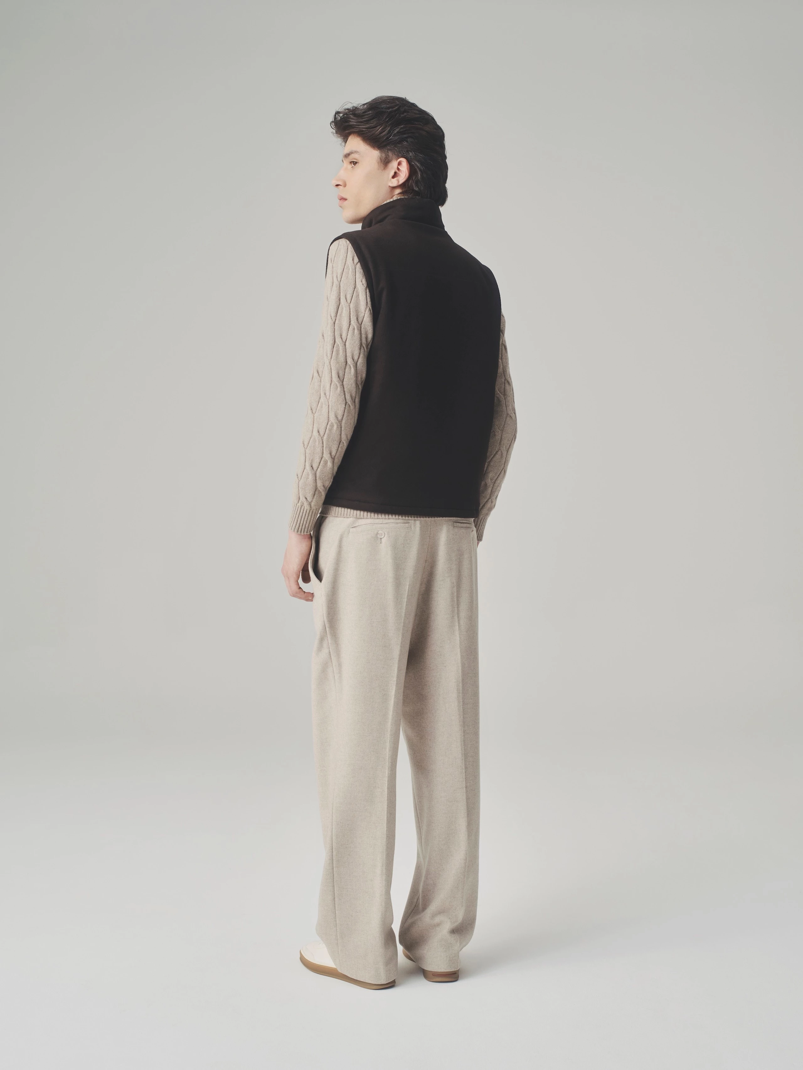Side view of a man in a studio wearing a brown bulletproof vest, beige cable knit sweater and wide wool trousers.