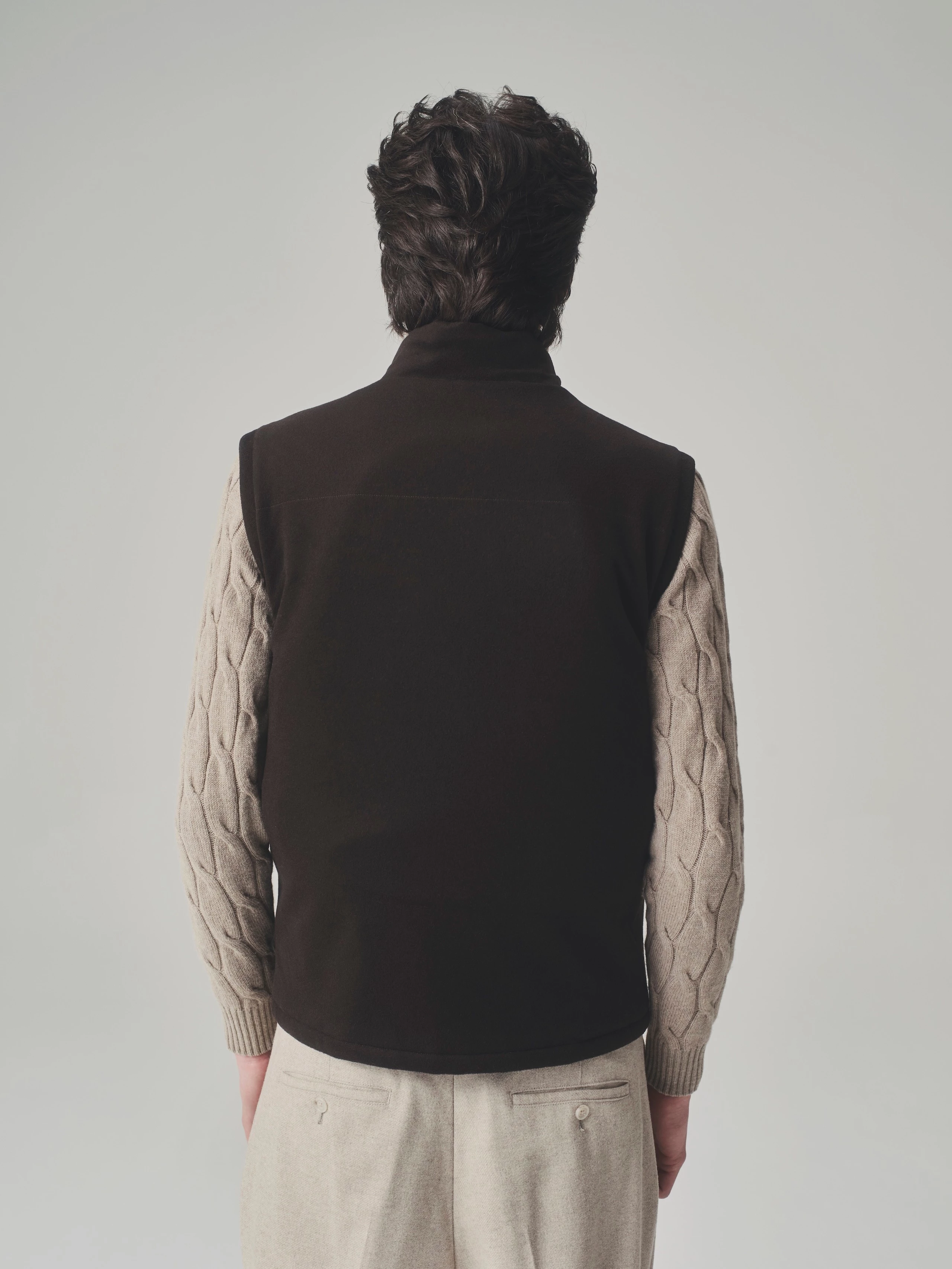 Back view of a man in a studio wearing a brown bulletproof vest, beige cable knit sweater and wool trousers.