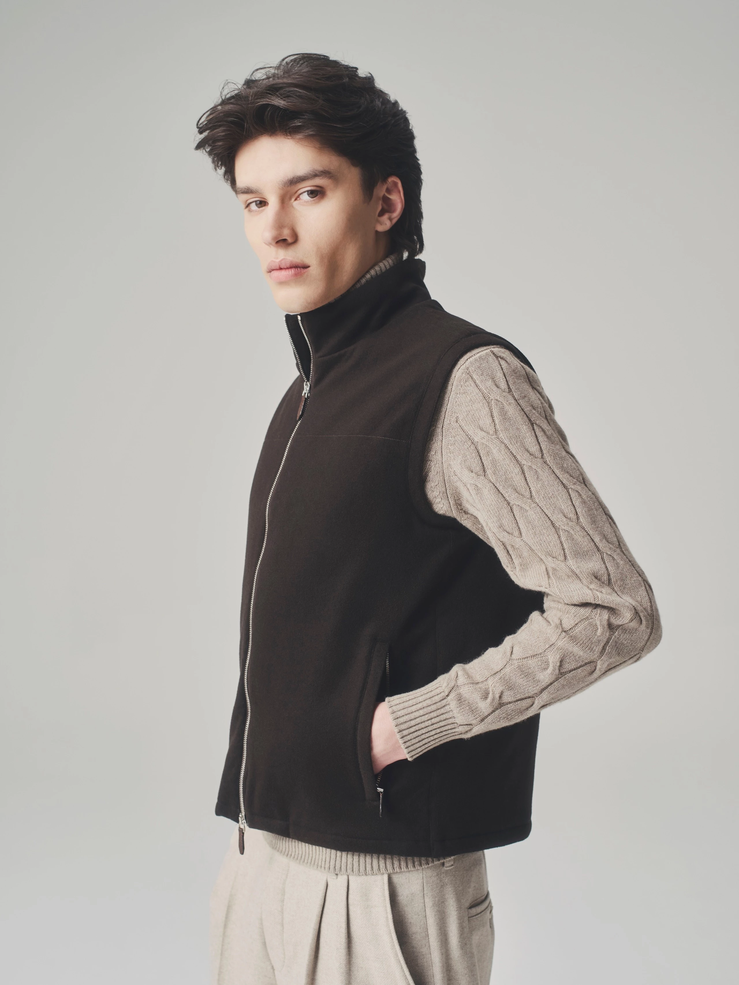 Portrait of a man in a studio wearing a brown bulletproof vest, beige cable knit sweater and wide wool trousers.