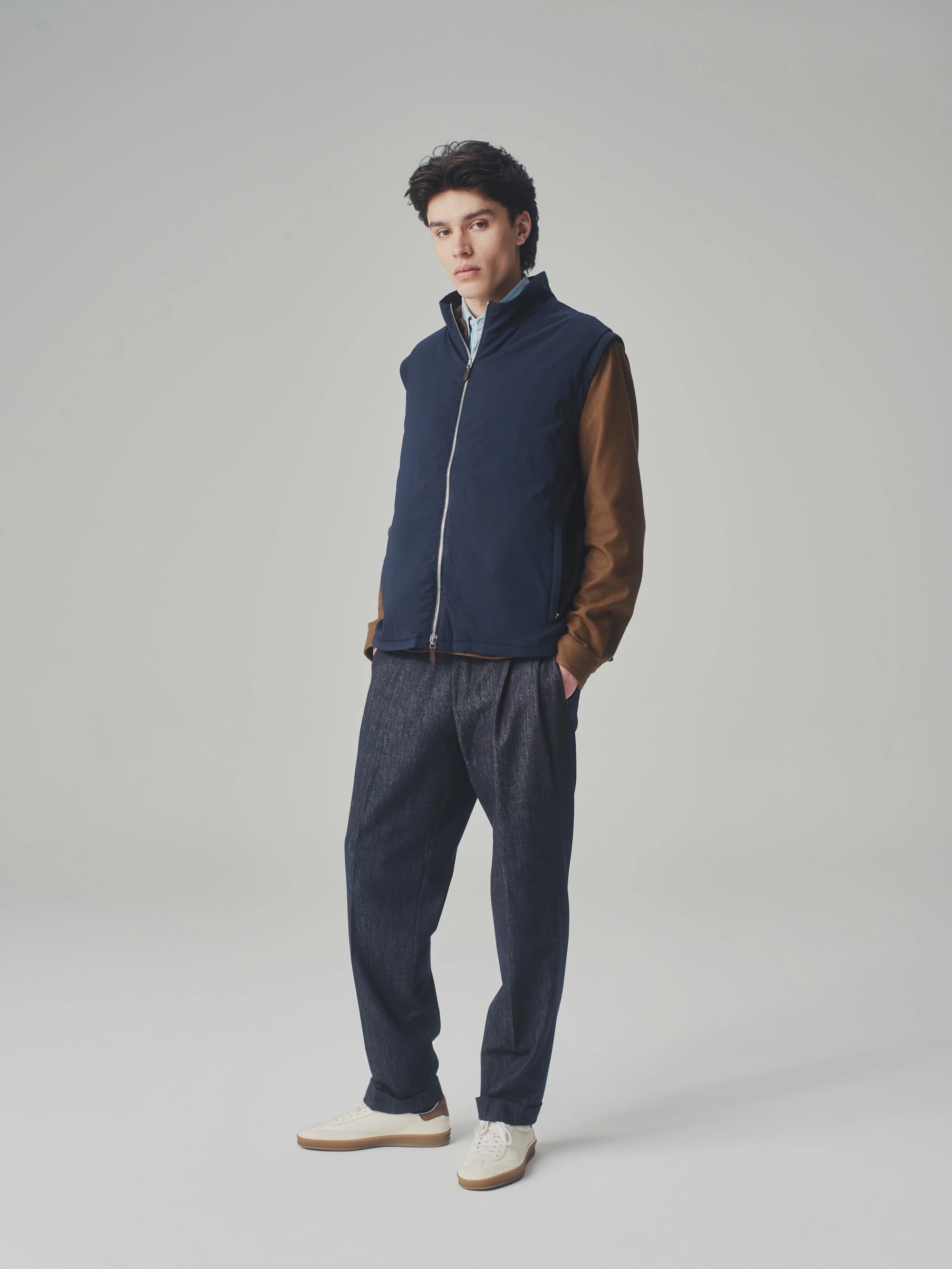 Man in a studio wearing a dark blue T-Way bulletproof vest over a brown jacket and light blue shirt with pleated trousers.