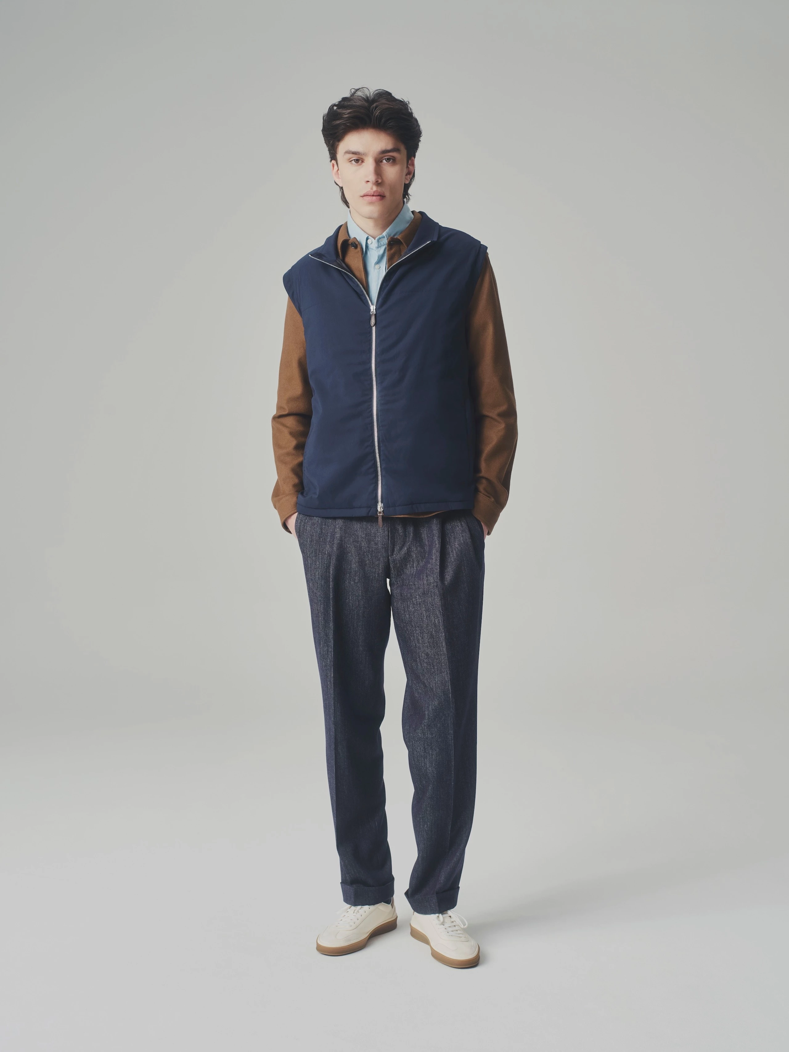 Man in a studio wearing a blue T-Way bulletproof vest over a brown jacket and light blue shirt with jeans and white sneaker.